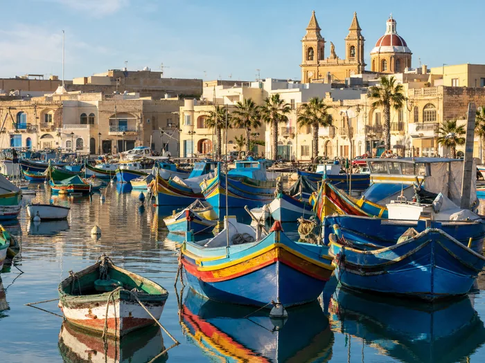 Painted luzzu fishing boats moored in a harbour lined with low buildings.